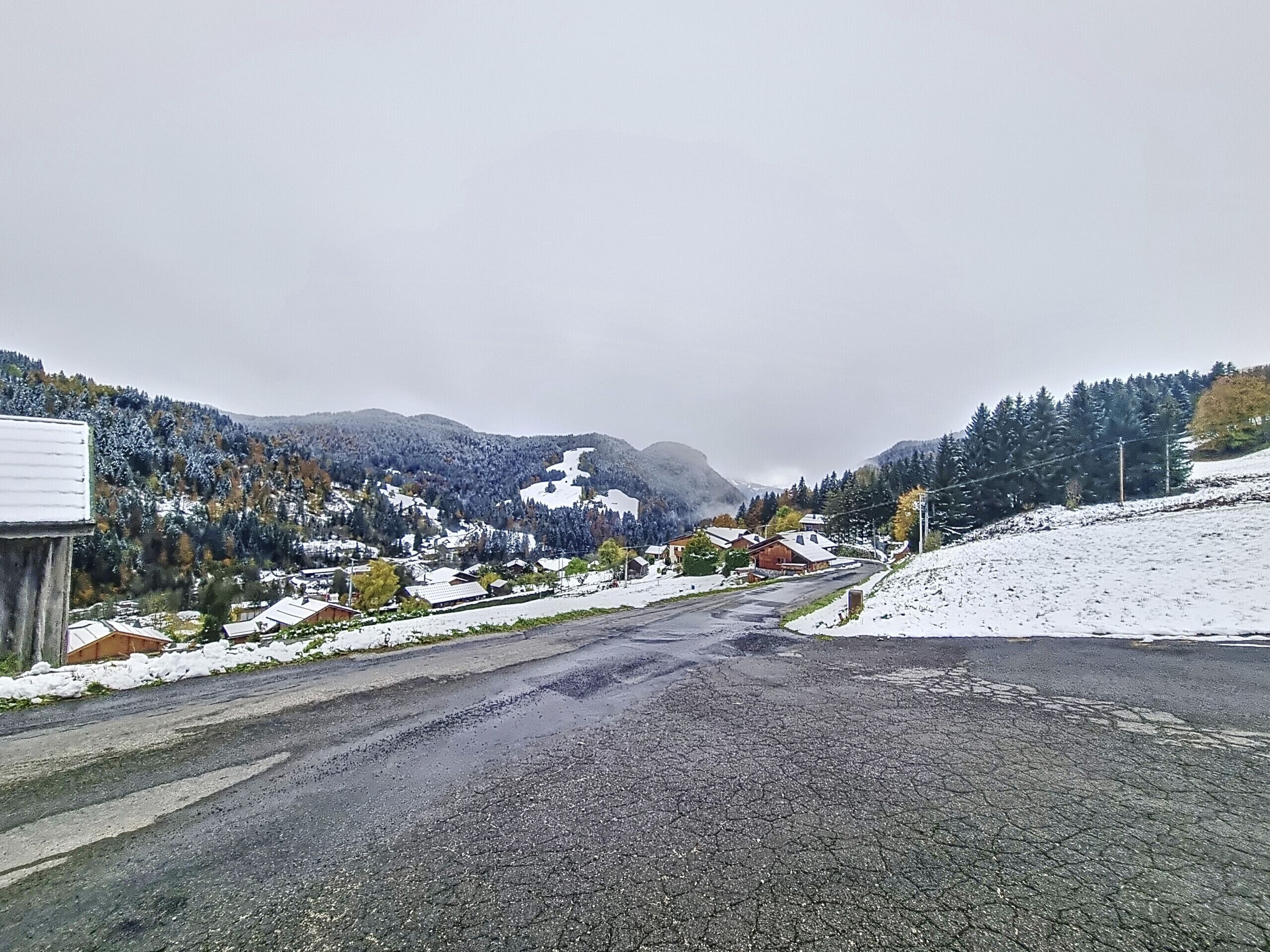 A rural road leading through a snowy landscape with houses and mountains in the background.