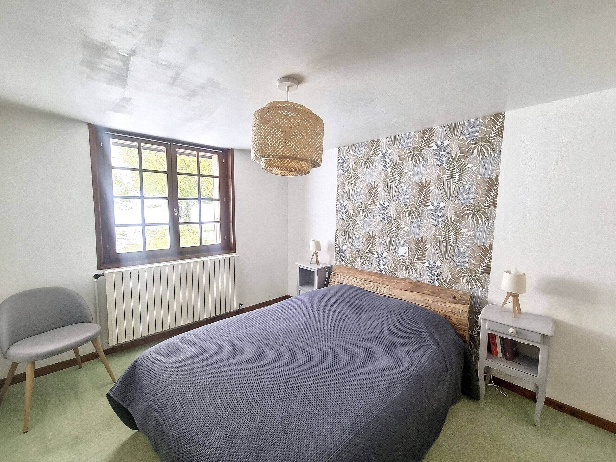 Een slaapkamer met een groot bed, een raam, een radiator, een stoel en een nachtkastje met een lamp.