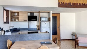 A compact kitchen with modern appliances, a dining area with a wooden table and chairs, and a living area with a sofa and a patterned wall. - Cornuts 2 - appartement -5pers (Les Gets)