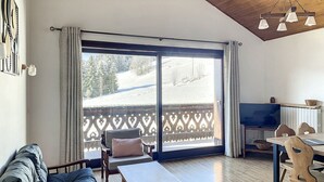 A cozy living room with a sofa, a wooden table, and a TV. A balcony with a wooden railing and a view of a snowy landscape is visible through the glass doors. - Cornuts 2 - appartement -5pers (Les Gets)