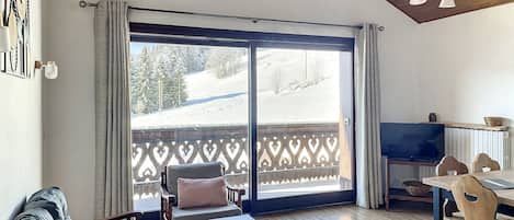 A cozy living room with a sofa, a wooden table, and a TV. A balcony with a wooden railing and a view of a snowy landscape is visible through the glass doors.