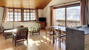 A cozy room with wooden floors, a dining area, a sofa, a TV, and large windows with curtains. - Cornuts 3 - appartement - 7pers (Les Gets)