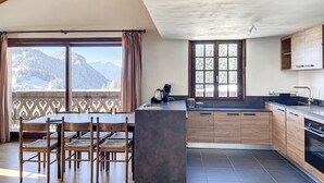 A modern kitchen with wooden cabinets, a large window with a mountain view, and a dining area with wooden chairs. - Cornuts 3 - appartement - 7pers (Les Gets)