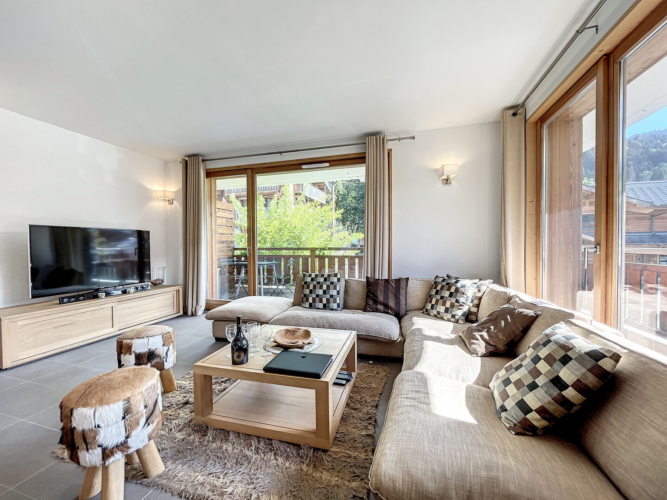 A modern living room with a large flat-screen TV, a sofa, a coffee table, and a view of a balcony with outdoor furniture.