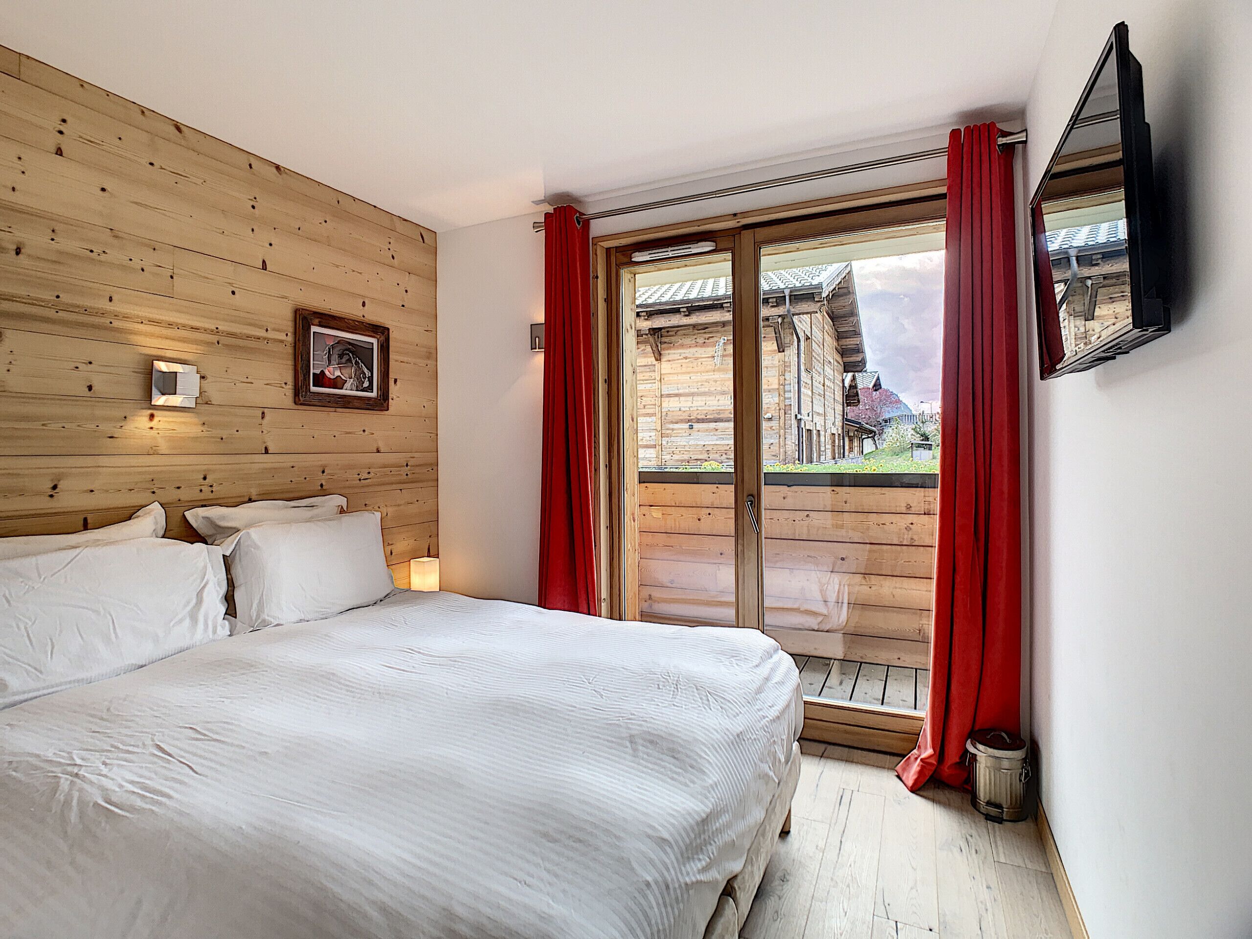 Une chambre avec un mur en bois, un lit, une fenêtre avec des rideaux rouges et une vue sur les bâtiments à l’extérieur.