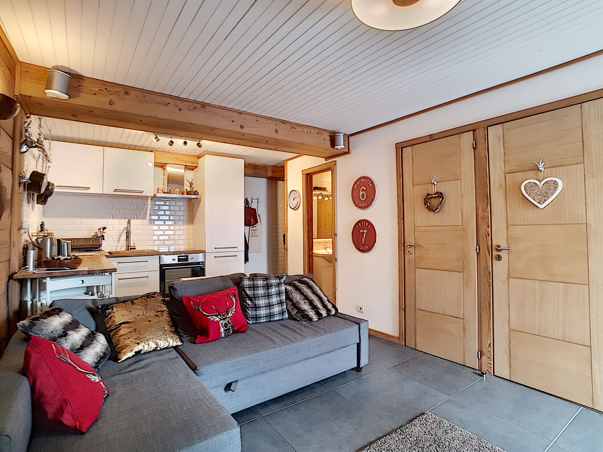 A modern living room with a grey sofa, wooden ceiling beams, a kitchen in the background, and a large wooden door.