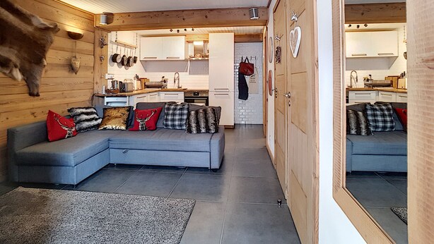 A modern interior with a grey sectional sofa, wooden walls, a kitchen with white cabinets, and a visible door leading to another room. - Montana - appartement - 6pers - proche centre (Les Gets)