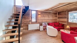 A wooden interior with a red sofa, a white chair, and a small table. A wooden staircase is visible on the left.