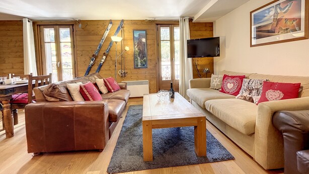 A cozy living room with a brown leather sofa, beige sofa, and a wooden coffee table. There is a TV mounted on the wall and a painting above the beige sofa.