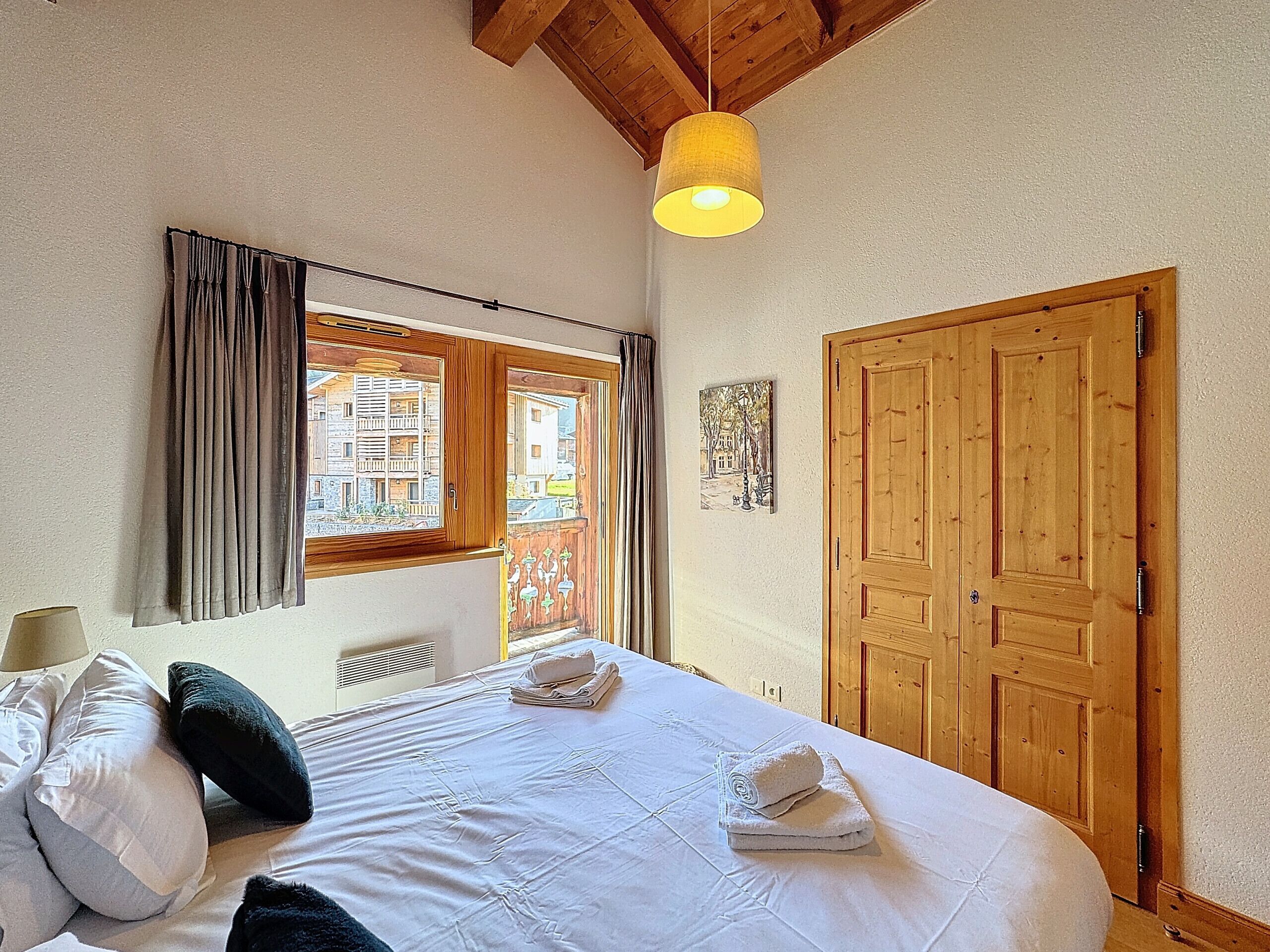 A bedroom with a bed, curtains, a wooden door, and a window with a view of buildings.