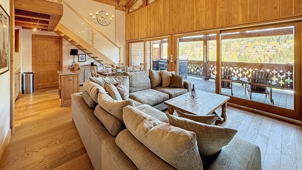 Ein geräumiges Wohnzimmer mit einer großen Couch, einem Essbereich mit Stühlen, einer Holztreppe und Blick auf eine gebirgige Landschaft durch große Fenster.