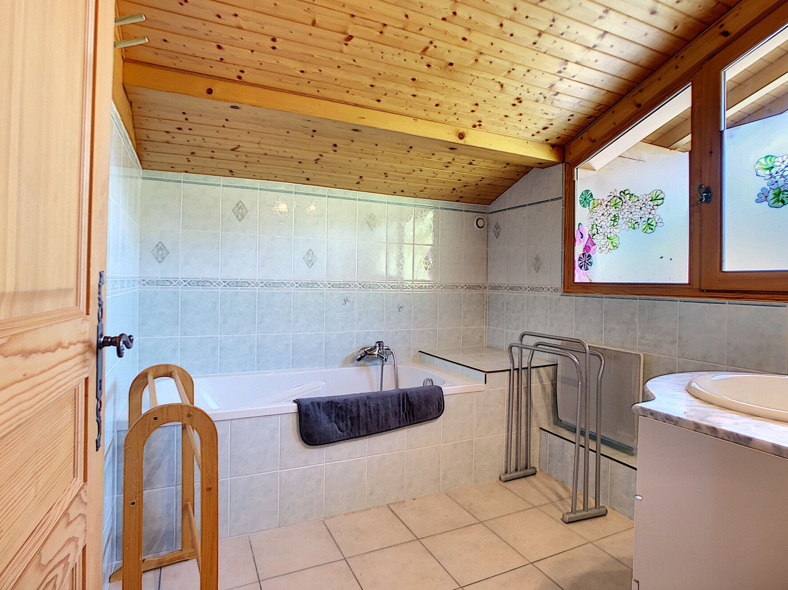 A bathroom with a wooden ceiling, a bathtub, a shower, a sink, and a window with floral decoration.
