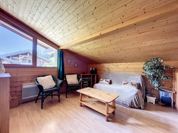 Une chambre confortable dans un chalet, avec un plafond en bois, un lit recouvert d’une literie à motifs, deux fauteuils en osier, une table en bois et une fenêtre donnant sur les bâtiments environnants.