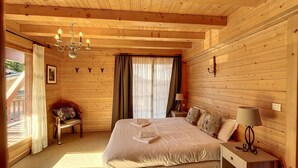 Ein Schlafzimmer einer Holzhütte mit einem Bett, einem Sessel, einem Nachttisch, einer Lampe und einem Fenster mit Vorhängen.