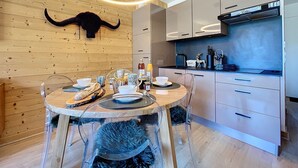 A modern kitchen with a round dining table, clear chairs, and a wooden wall with a mounted animal head. - Les 3 Sapins - appartement4pers - centre station (Les Gets)