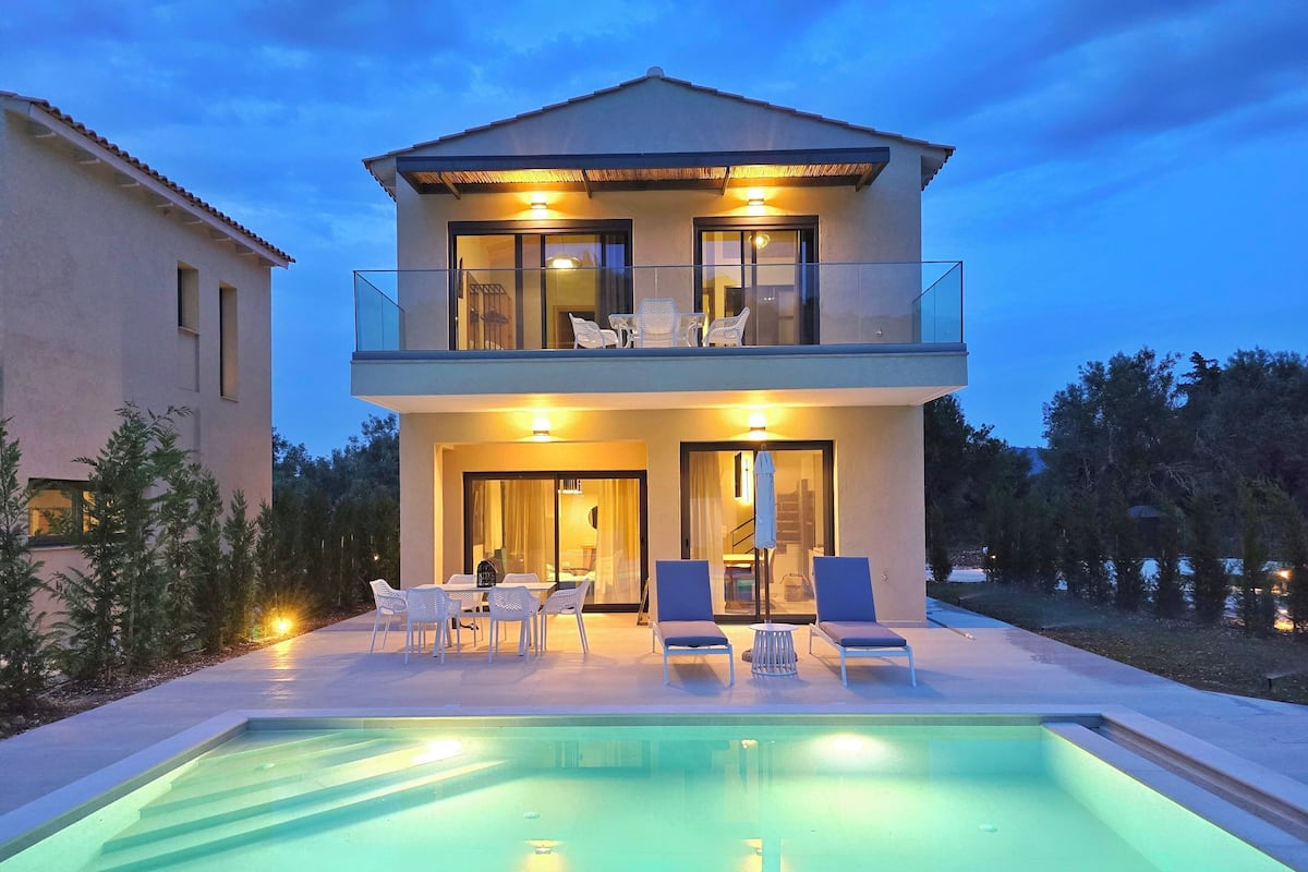 A two-story modern house with a swimming pool, outdoor seating, and a well-lit entrance.