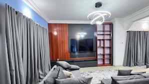 Een moderne woonkamer met een grote flatscreen-tv, grijze gordijnen, een wand van houten panelen en een inbouwboekenrek.