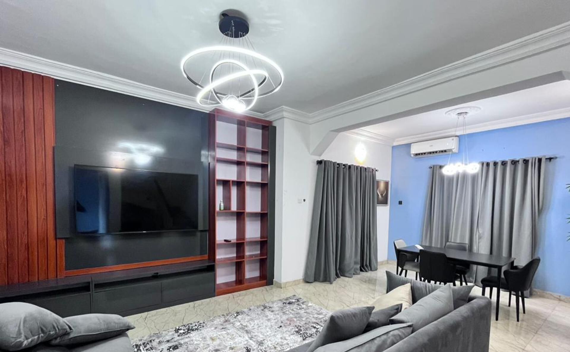 Una sala de estar moderna con un televisor de pantalla plana, una estantería empotrada, cortinas grises, un comedor con mesa y sillas, y un equipo de aire acondicionado.