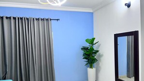 Een kamer met een blauwe wand, grijze gordijnen, een witte plant in een witte pot en een spiegel met een zwarte lijst.