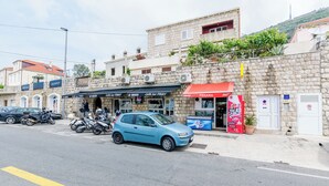 A street view with a blue car parked in front of a building with multiple shops, including a café and a juice bar. - Apartment Bim (Dubrovnik)