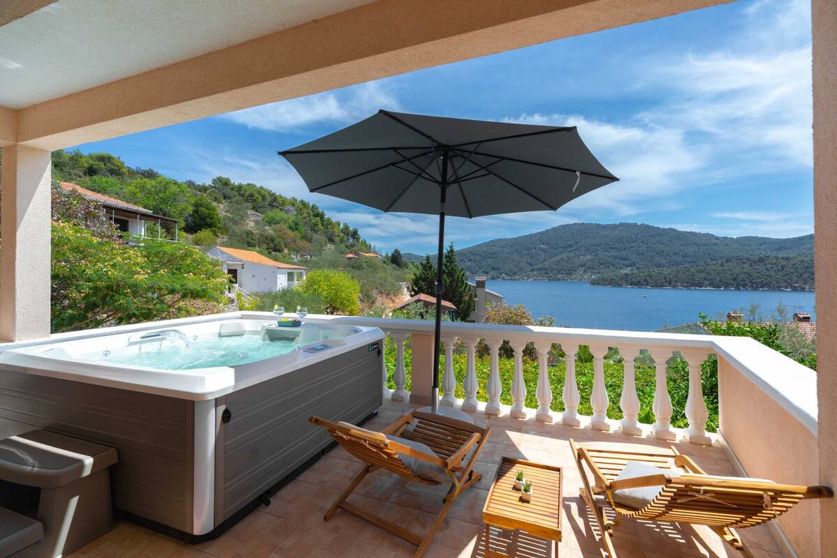 Un balcon doté d’un jacuzzi, de chaises en bois et d’une table, offrant une vue sur un lac et des collines.