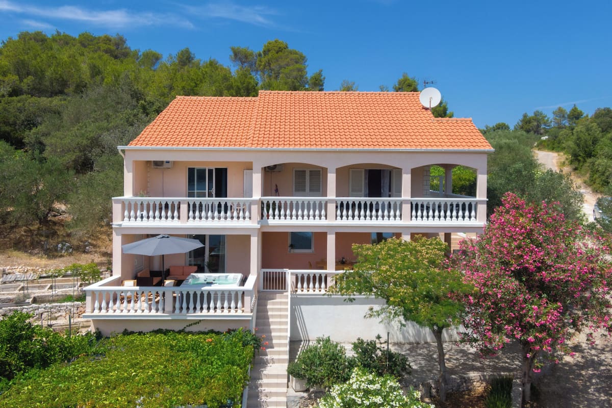 Un bâtiment à deux étages, doté d’un toit recouvert de tuiles rouges, d’un balcon avec des balustrades blanches et d’un jardin planté de fleurs.