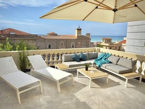A balcony with white furniture, a beige umbrella, and a view of the sea and buildings.