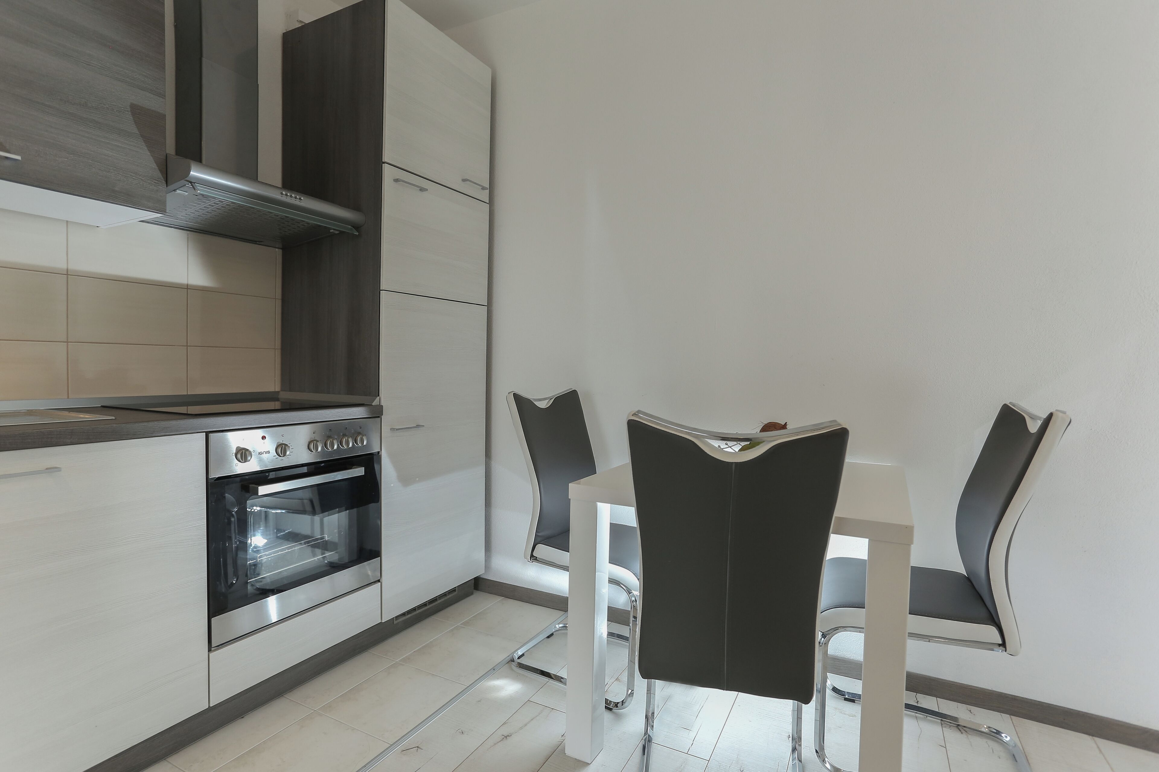 A modern kitchen with a built-in oven, a stainless steel range hood, and a dining area with a white table and black chairs.