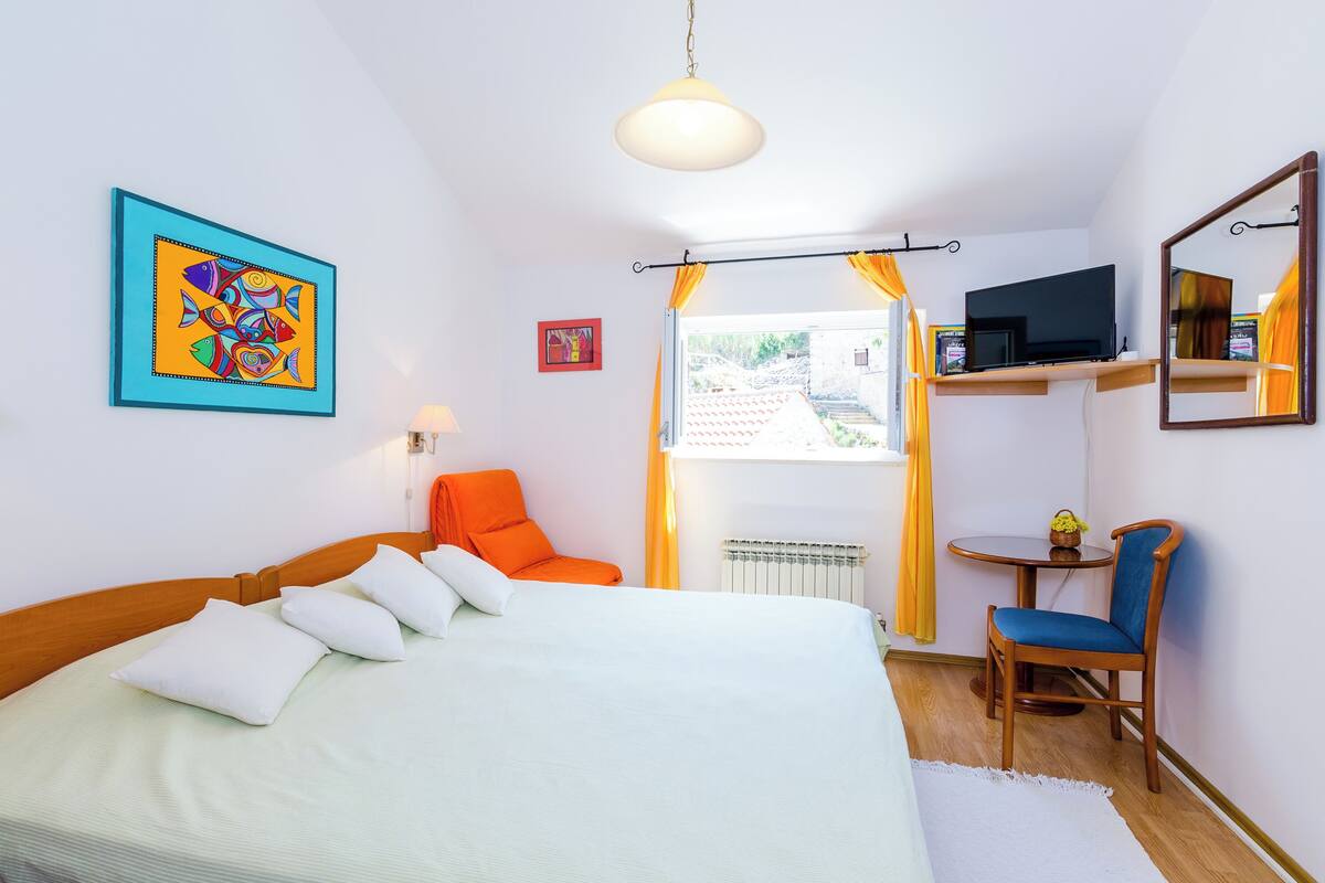 A bedroom with a bed, a chair, a TV on a shelf, and a painting on the wall.