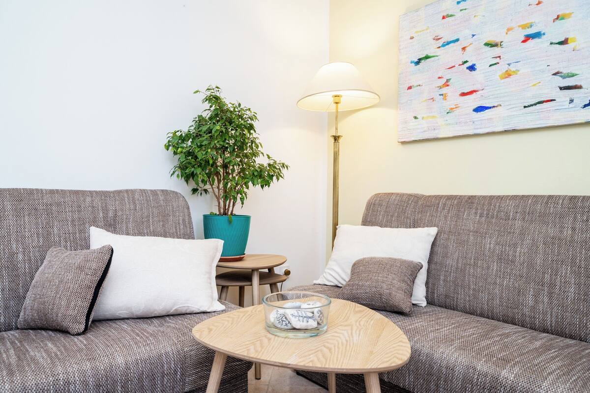 A modern living room with two grey sofas, a round wooden coffee table, a teal potted plant, a floor lamp, and a colorful abstract painting on the wall.