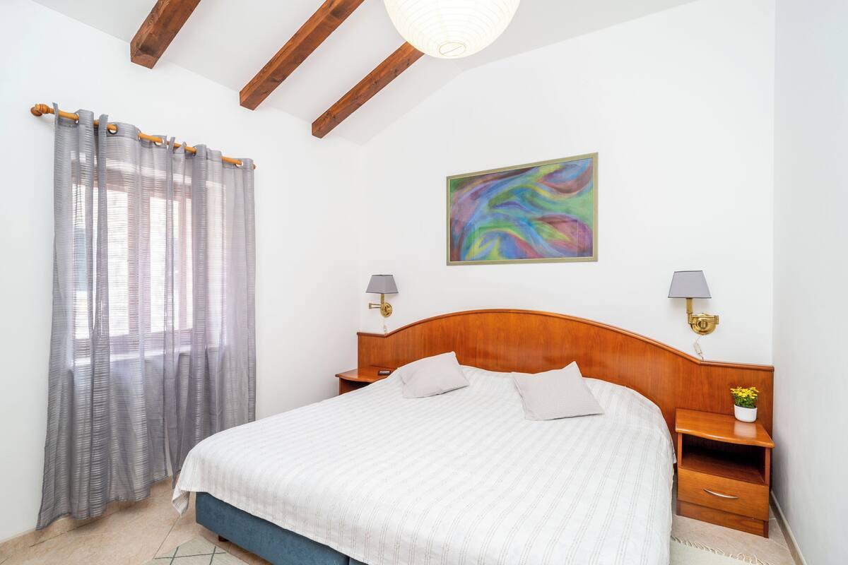 A bedroom with a wooden headboard, a bed with white bedding, two bedside tables with lamps, a colorful abstract painting on the wall, and sheer curtains on the window.