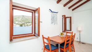 A dining area with a wooden table and chairs, a window with an open shutter, and a view of a body of water and hills. - Art Apartment Seasoul (Dubrovnik)