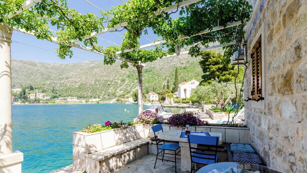 A stone patio with a table and chairs overlooking a body of water, surrounded by greenery and a mountainous backdrop. - Art Apartment Seasoul (Dubrovnik)