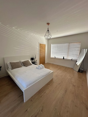 A modern bedroom with a large bed, a wooden floor, a ceiling light fixture, and a window with blinds.