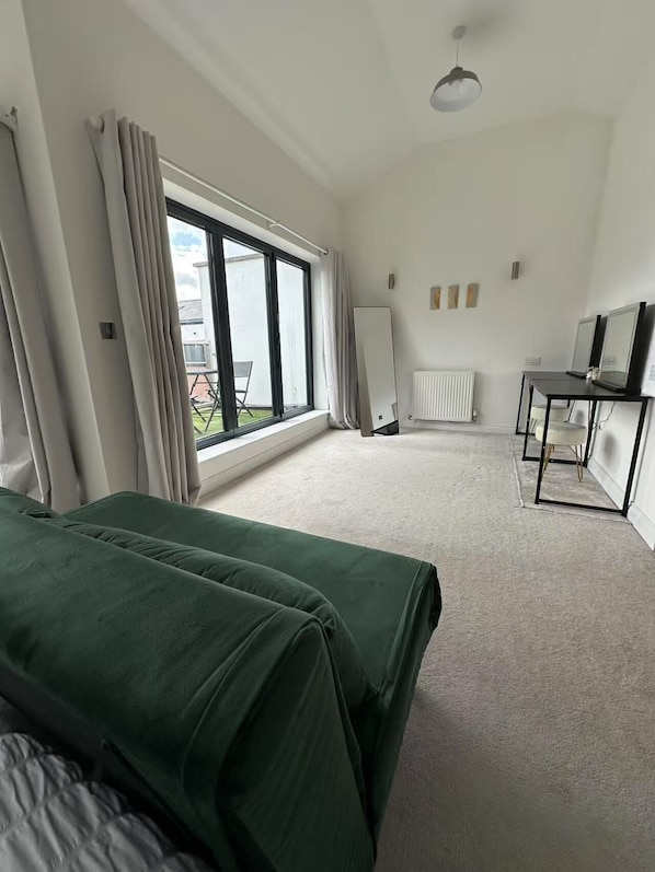 A spacious room with a large window, a bed, a desk with a mirror, and a radiator.