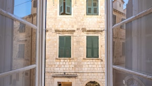 Visão pela janela de um edifício de pedra com três venezianas verdes, uma porta e uma pequena loja no nível da rua.