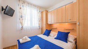 Ein Schlafzimmer mit Bett, einem flachbildfernseher an der Wand, Holzausbau und einem Fenster mit leichtem Vorhang.