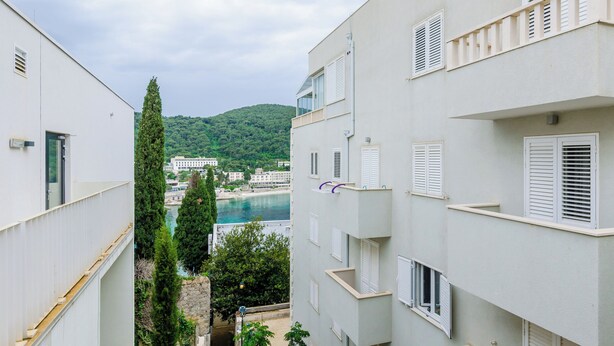 A narrow street between two white buildings with shutters, overlooking a body of water and a hillside. - Grand Apartment (Dubrovnik)