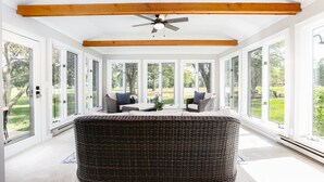 Una sala de estar con ventanales amplios, ventilador de techo, mobiliario de mimbre y vistas a un jardín.