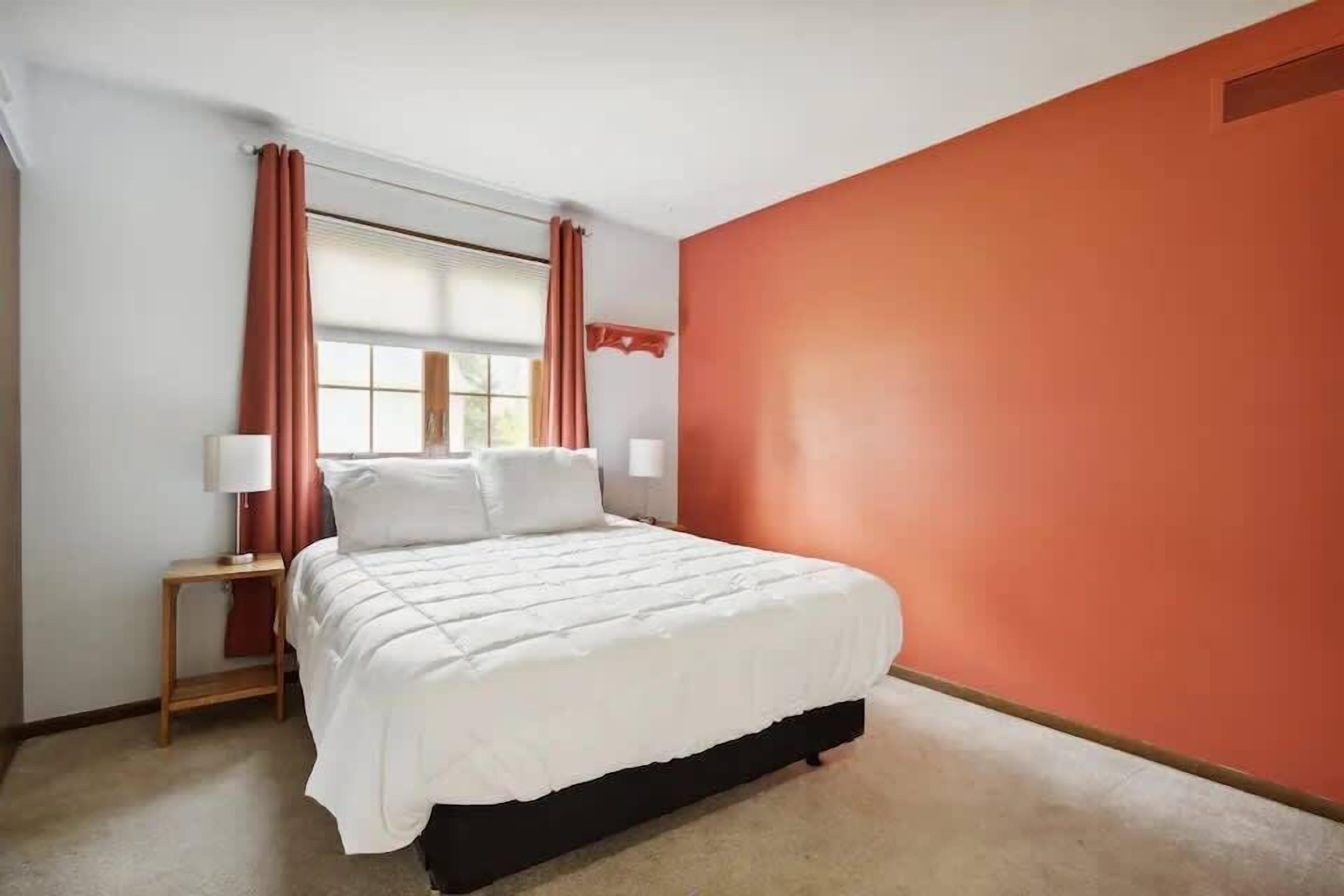 Ein Schlafzimmer mit einem Bett, zwei Nachttischen, einem Fenster mit Vorhängen und einer orangefarbenen Wand.