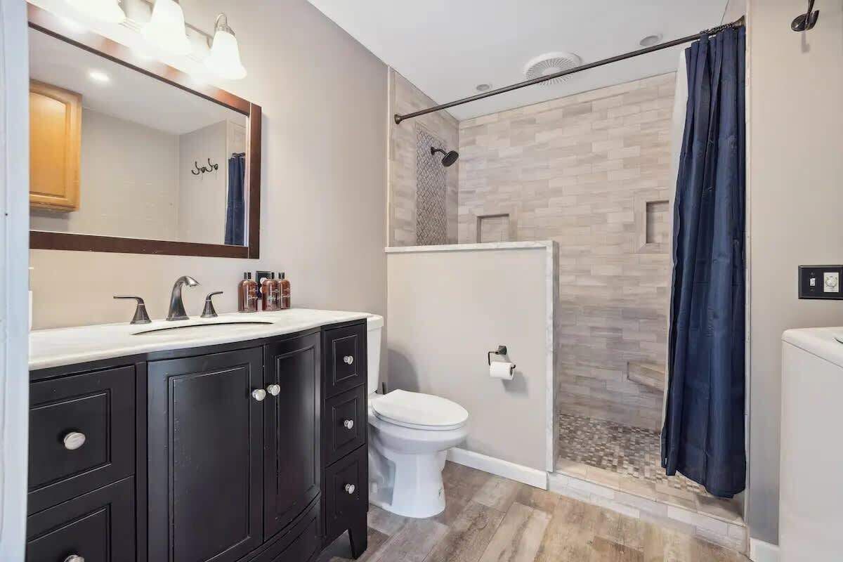 Un baño moderno con ducha, inodoro y tocador con muebles oscuros y un espejo.