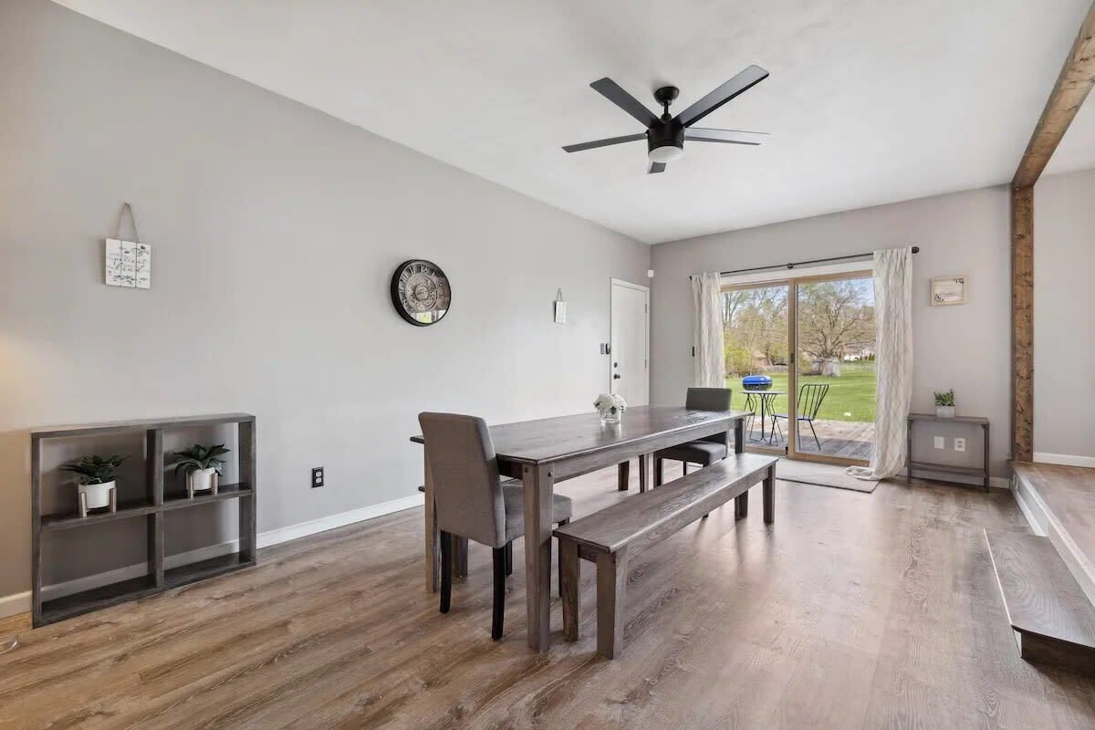 Ein Essbereich mit Holztisch, Bänken und Deckenventilator. Eine Glastür führt zu einer Terrasse mit Gartenmöbeln.
