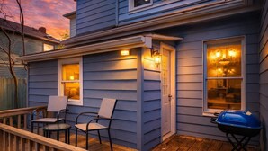 Una casa con terraza de madera, dos sillas, una parrilla y luces exteriores.