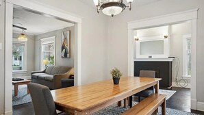 Un comedor amplio con una mesa de madera, bancos y una planta en maceta. Se observa un sofá y una chimenea al fondo.