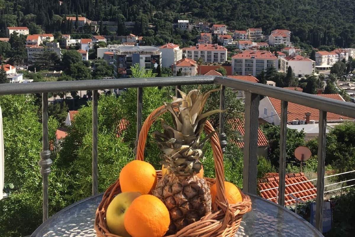 A wicker basket with fruits on a balcony overlooking a town with buildings and a hill.