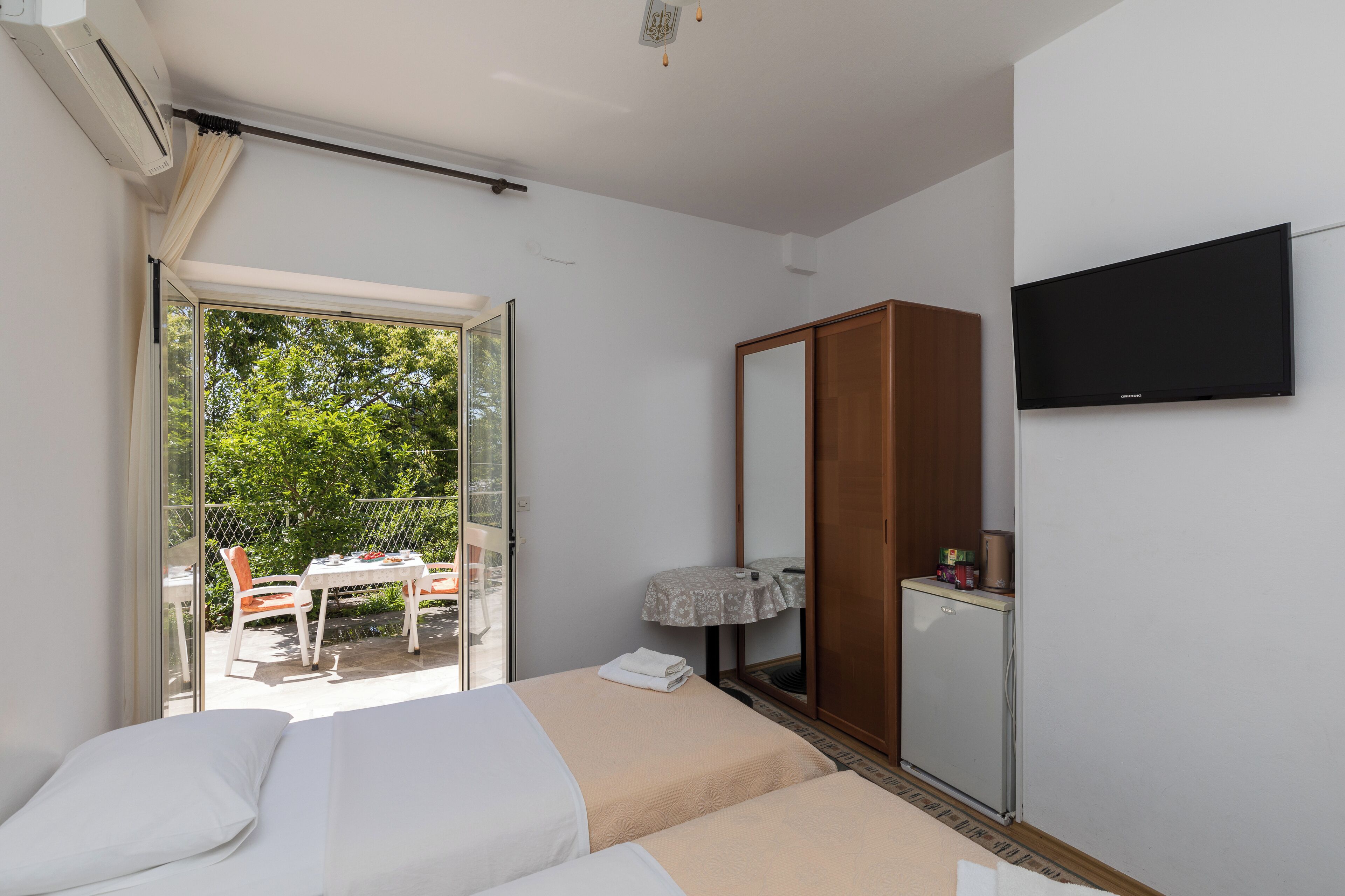 Ein Hotelzimmer mit Bett, Fernseher, Kleiderschrank, einem kleinen Tisch mit Tischdecke und Blick auf einen Außenbereich mit Tisch und Stühlen.