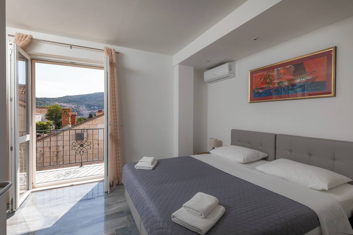 A bedroom with a large bed, a painting on the wall, and a balcony with a view of the city.