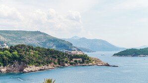 Paisatge costaner amb un edifici històric en un turó, una massa d'aigua i muntanyes a la distància.