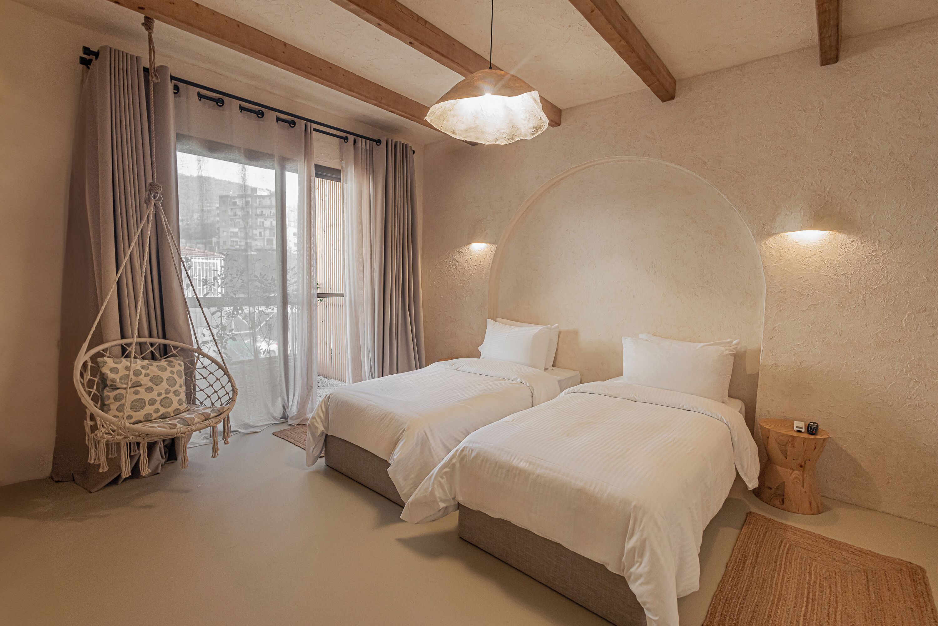 A bedroom with two beds, a hanging chair, a small wooden side table, and a large window with curtains.