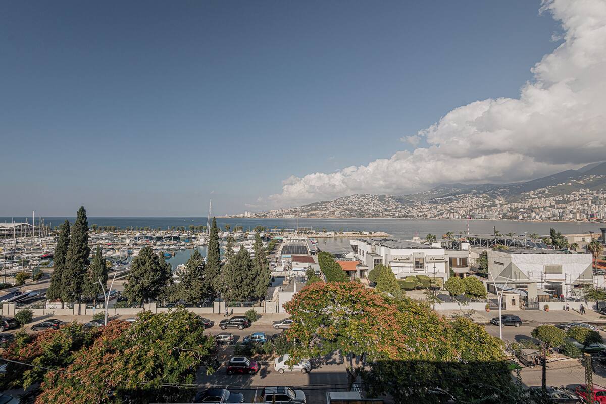 A marina with boats, a parking area, and buildings with a view of the sea and mountains.